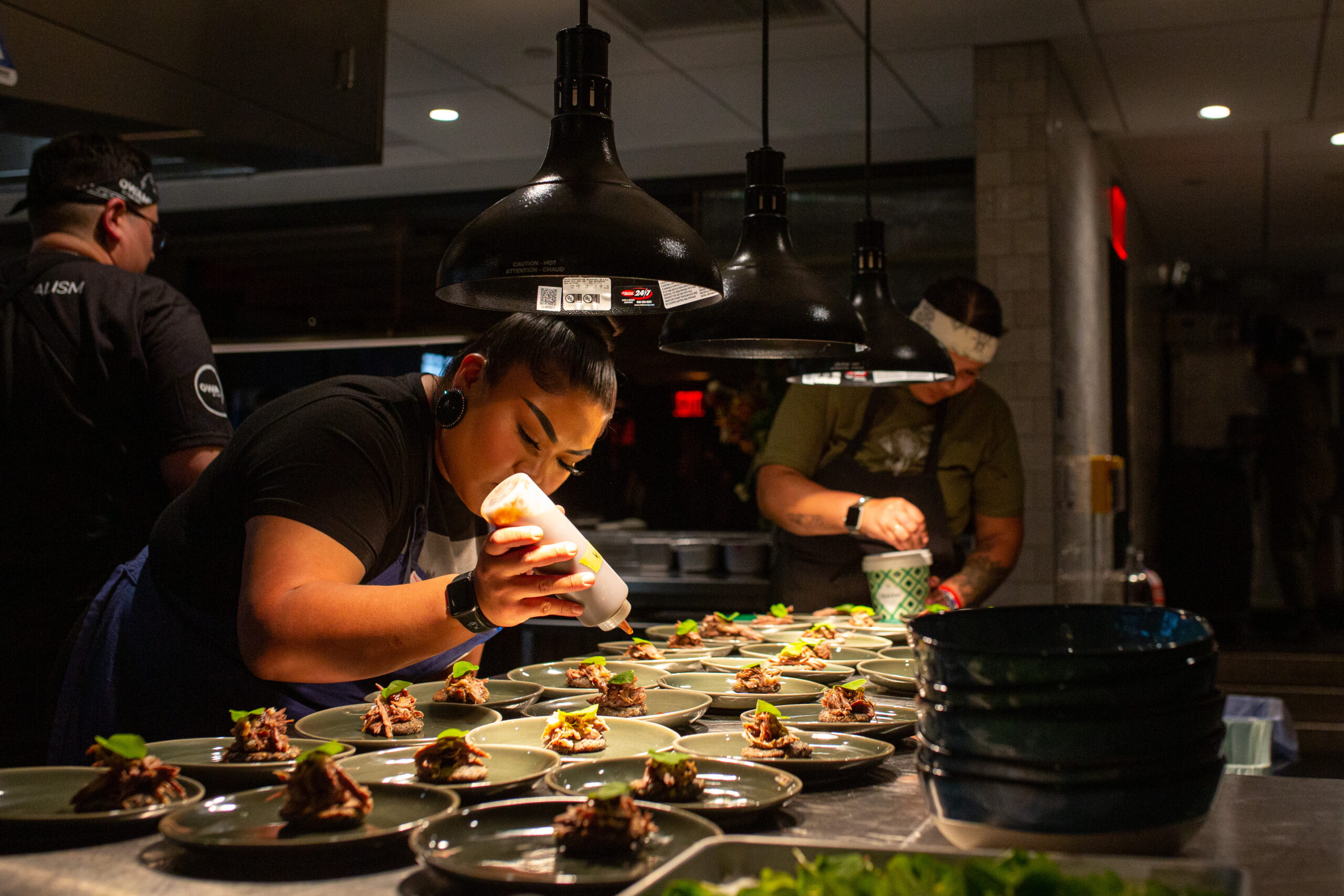 Chefs in a kitchen put the final touches on a row of dishes, all garnished with a leafy green.