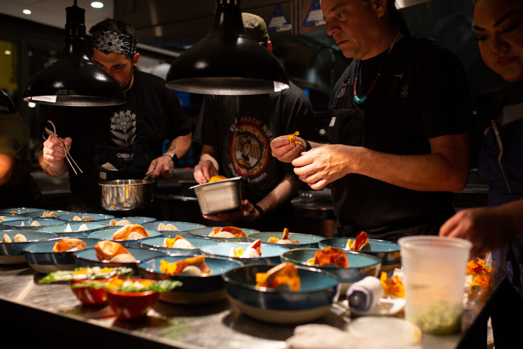 A team of chefs prepare three rows of plated dishes, lit from above by spotlights.
