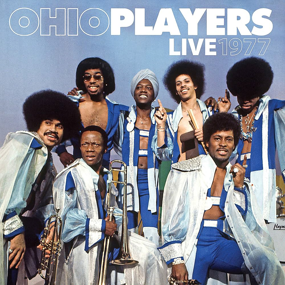 An old record cover that reads "Ohio Players Live 1977." There are seven people in the photo used on the cover and some of them are holding trumpets and drumsticks They are all stylishly dressed in clothing made of bright blue, silver, and white fabrics. Some of the members have natural Afro hairstyles, while one is wearing a turban.