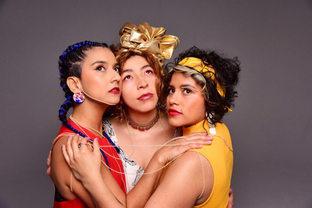 Four women of medium skin tone wearing brightly colored outfits in front of a gray background, embracing and looking upwards.