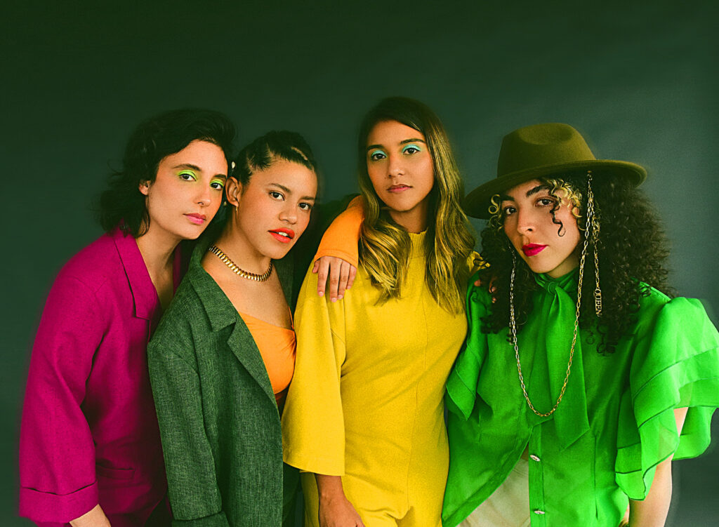Four women of medium skin tone wearing brightly colored blouses in front of a green background.