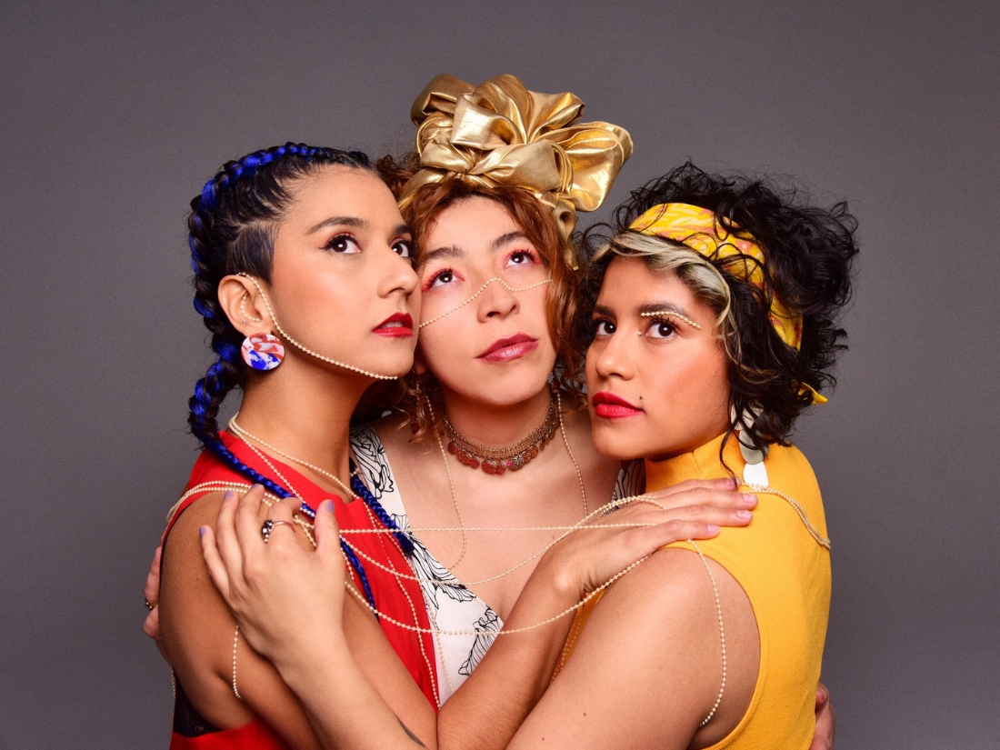 Four women of medium skin tone wearing brightly colored outfits in front of a gray background, embracing and looking upwards.