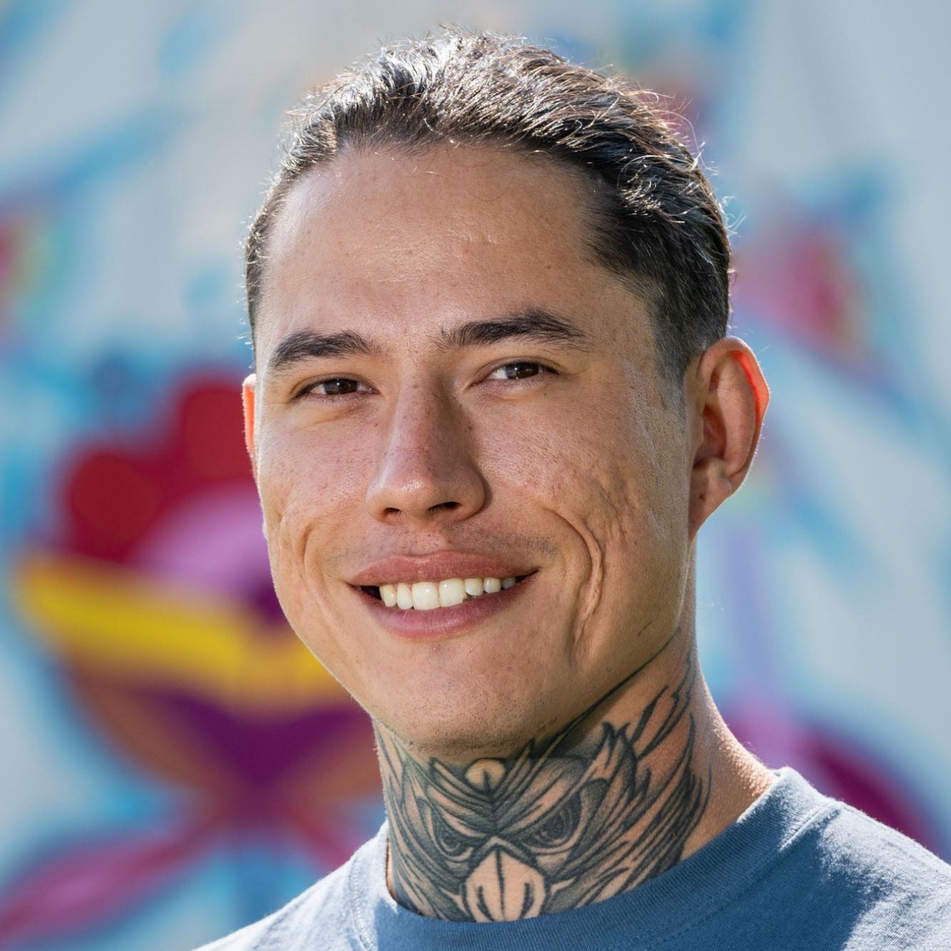 Headshot of a person with medium-light skintone and a neck tattoo wearing a t-shirt in front of a colorful mural.