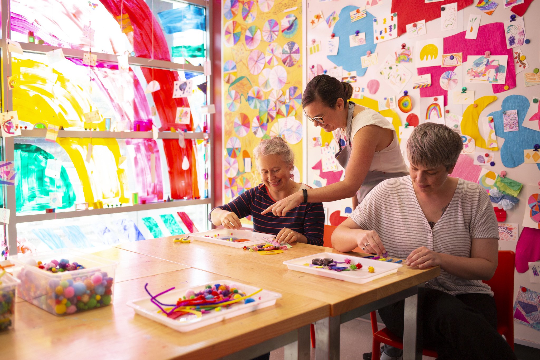 A person in an apron directs two people working on a craft with pom poms in a room decorated floor-to-ceiling in colorful art.