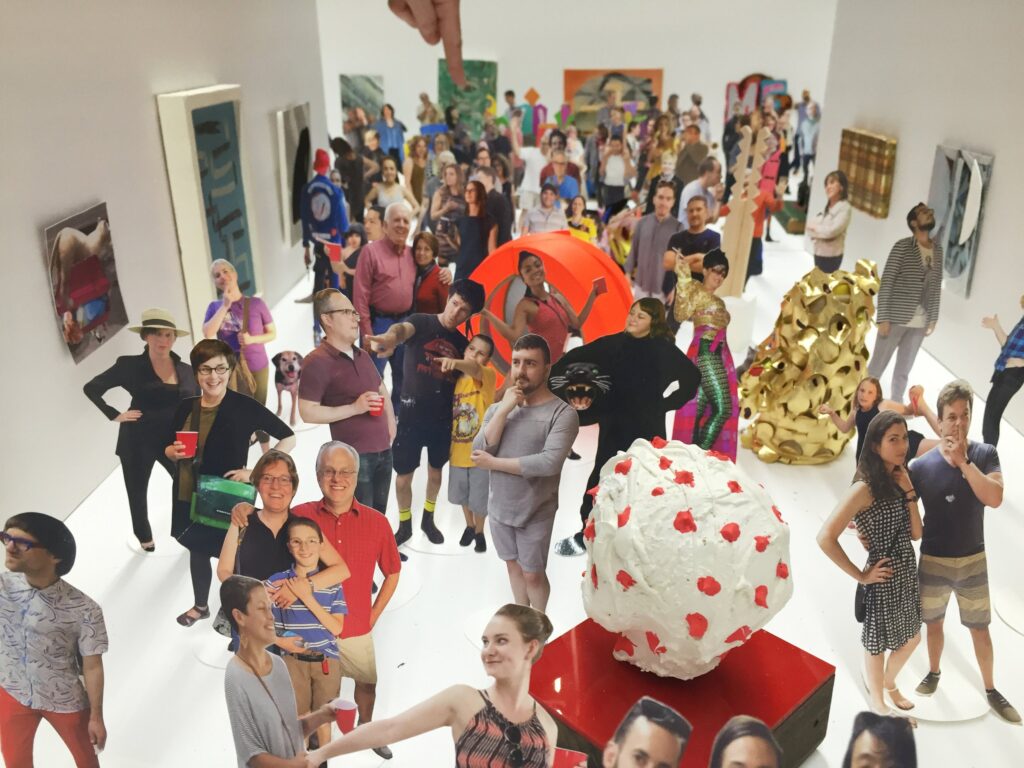 A white box filled with miniature cutouts of people and scaled down art pieces to resemble a model of a gallery space. A person's finger is seen at the top pointing to one of the figures in the back.