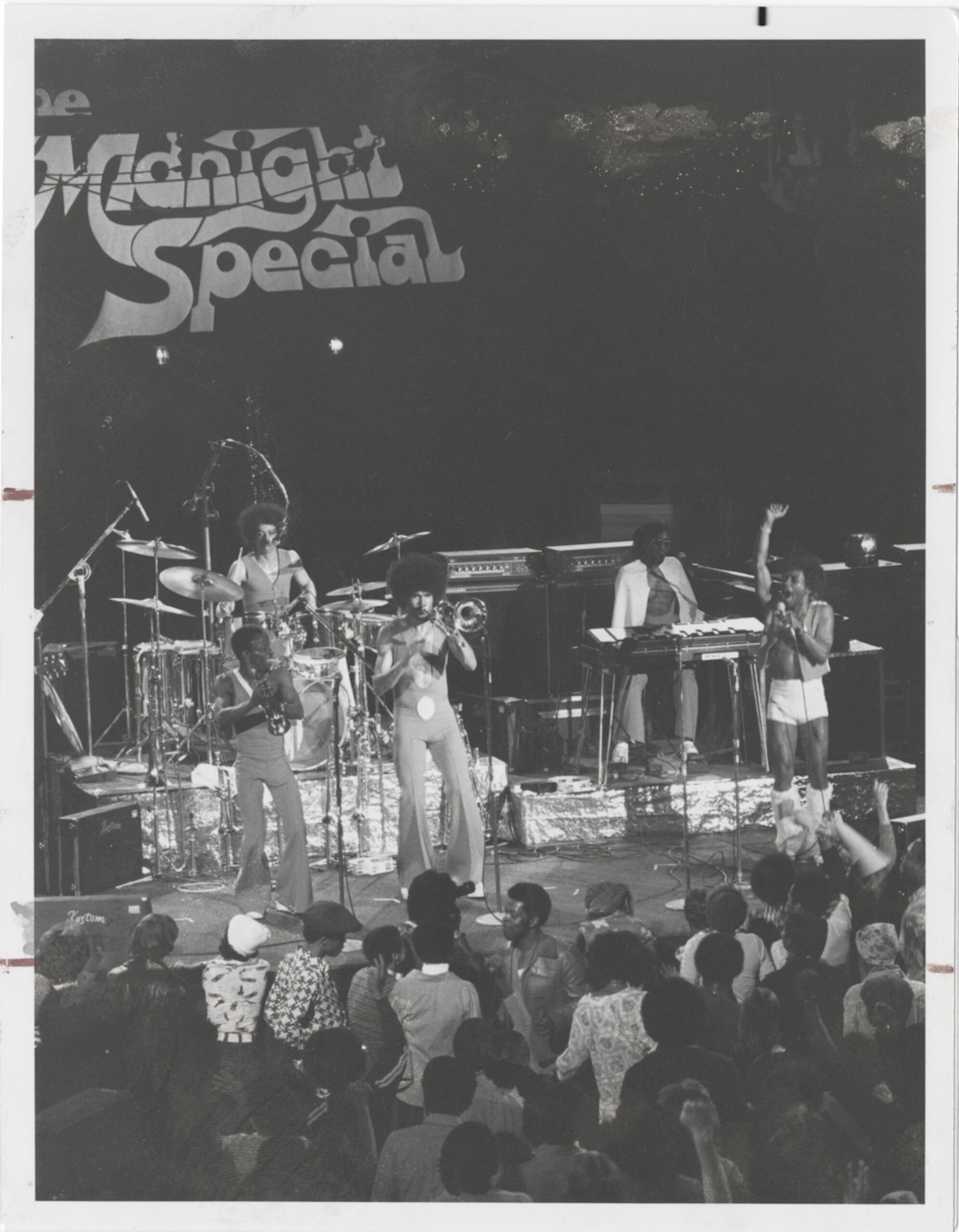 A black and white photo of a band playing instruments and singing into microphones on stage in front of an audience. The band is stylishly dressed in bellbottom pants and some of the members have natural Afro hairstyles. In the background, there are letters that read "Midnight Special"