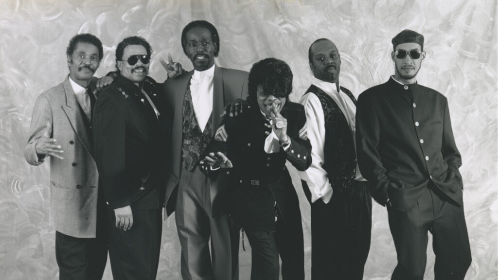 A black and white photo of six people standing close to each other, posing for their band photo. They are wearing different styles of suits and have different hairstyles. One of them, centered in the photo, is a little hunched over, gesturing with their hand over their mouth and grinning.