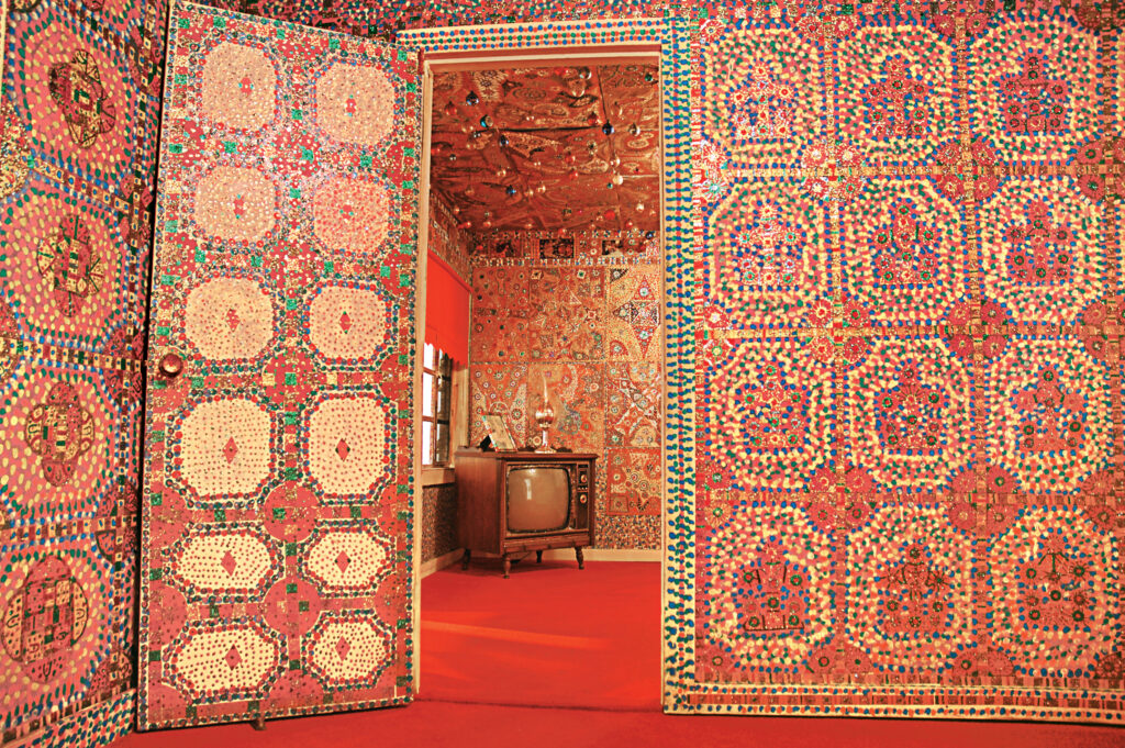 A room embellished, ceiling to floor, with multicolor rhinestone in varying patterns. A door leads to another room with similar aesthetics and a wooden box television.