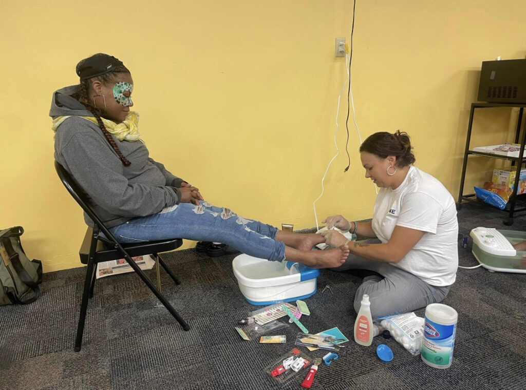 A person wearing an eye mask sitting in a folding chair while another person gives them a pedicure.