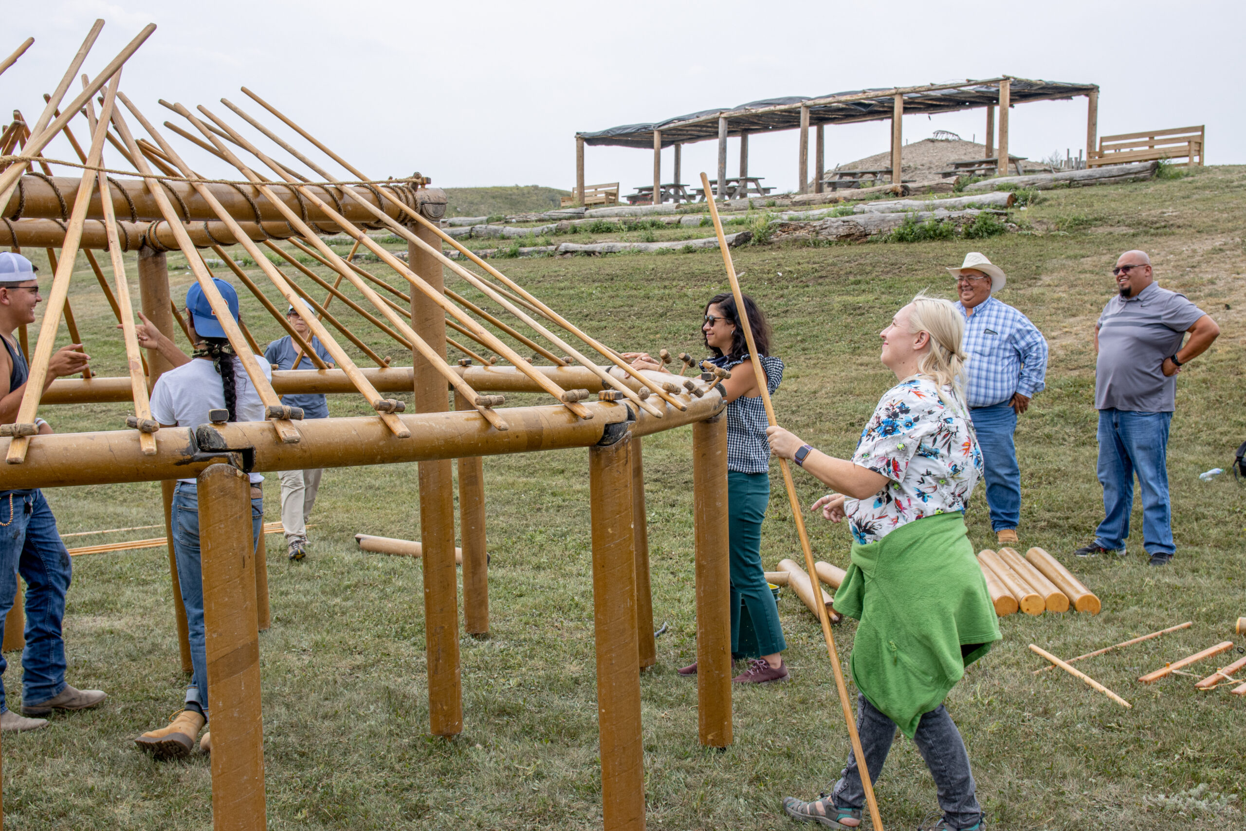 A group of people hold wooden building materials of varying sizes and shapes to collectively build a circular outdoor shelter.