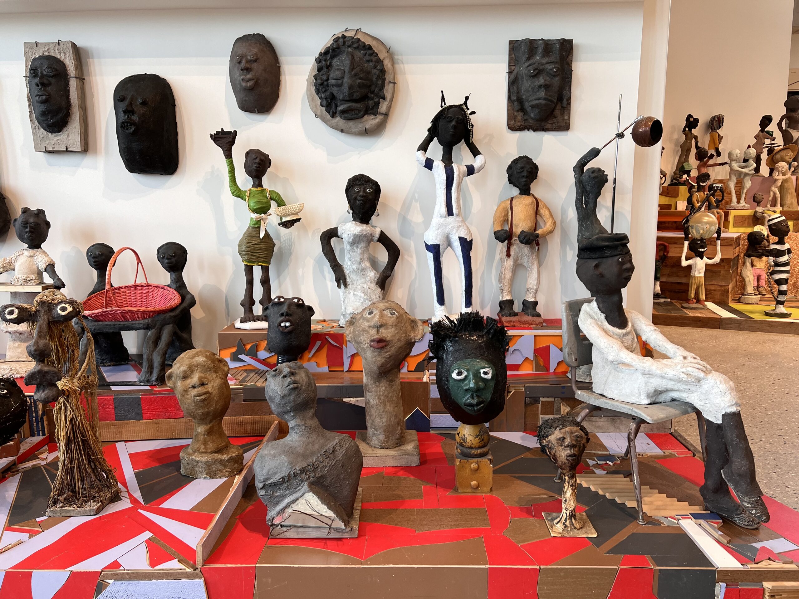 A collection of sculptural works depicting figures with dark skin tone. Some are mounted on the wall and others are displayed on a table-like surface with a patterned covering.