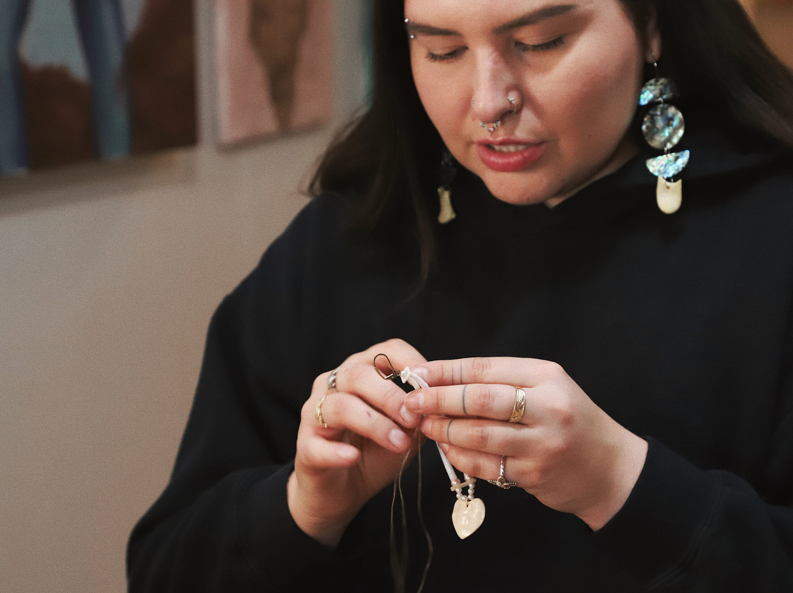 A person of medium skin tone with dark long hair, wearing a black hooded sweatshirt, works with their hands to make an earring.