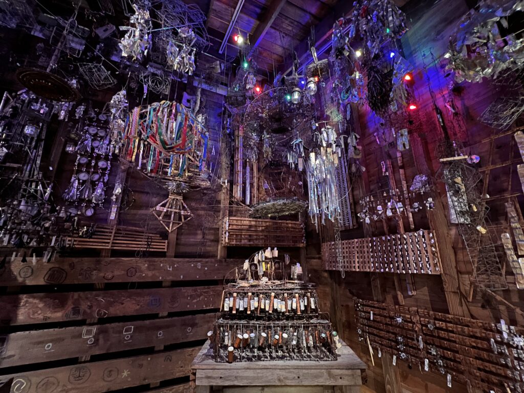 A low-lit room with sculptural works made of silvery tinsel and other fringe-like elements hanging from the ceiling. The worn wooden walls have drawn symbols and motifs.