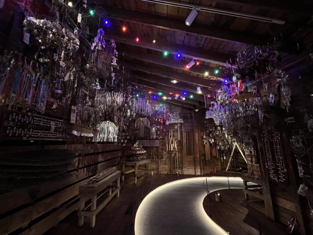 A low-lit room with sculptural works made of silvery tinsel and other fringe-like elements hanging from the ceiling. The worn wooden walls have drawn symbols and motifs.