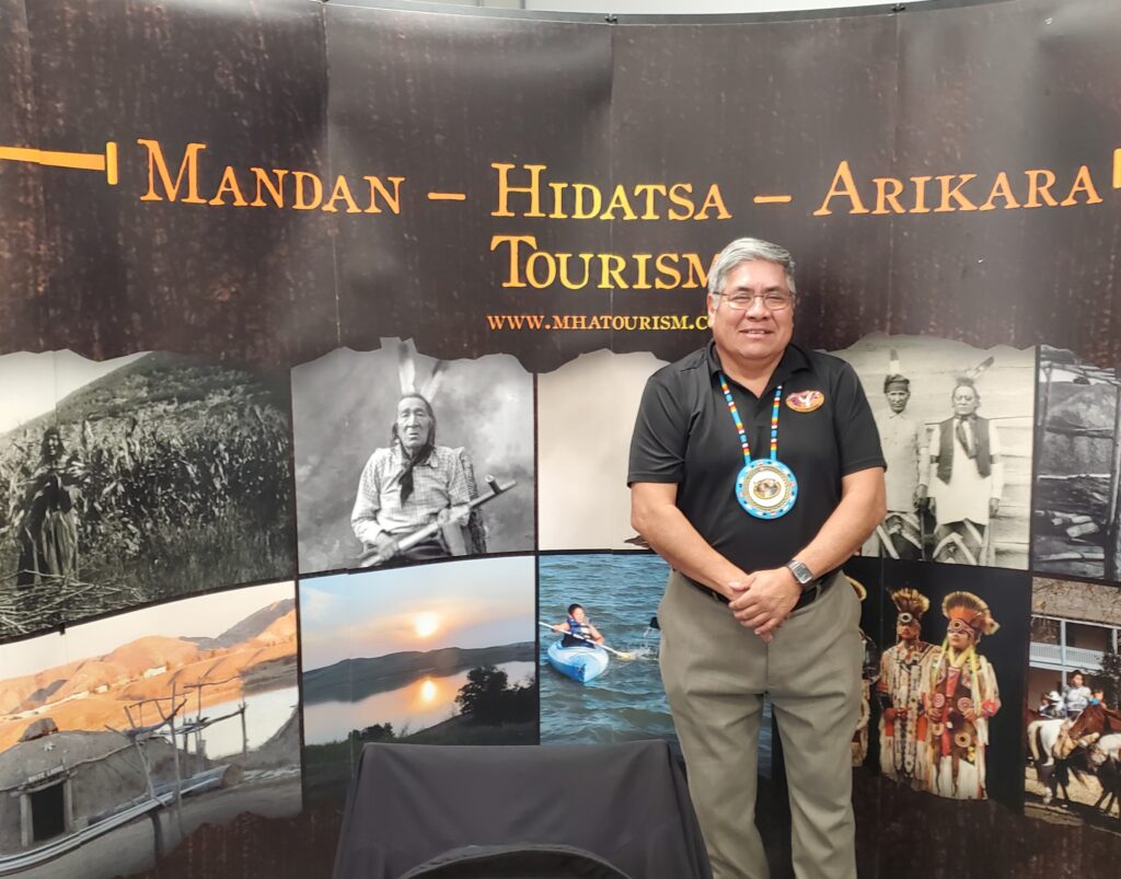 A person of medium skin tone smiles and poses for the camera. They have short grey hair and wearing a black polo shirt and grey trousers, and have a Native American beaded necklace. They are standing against a backdrop that has images of Native American peoples and large text that reads "Mandan - Hidatsa - Arikara Tourism'.