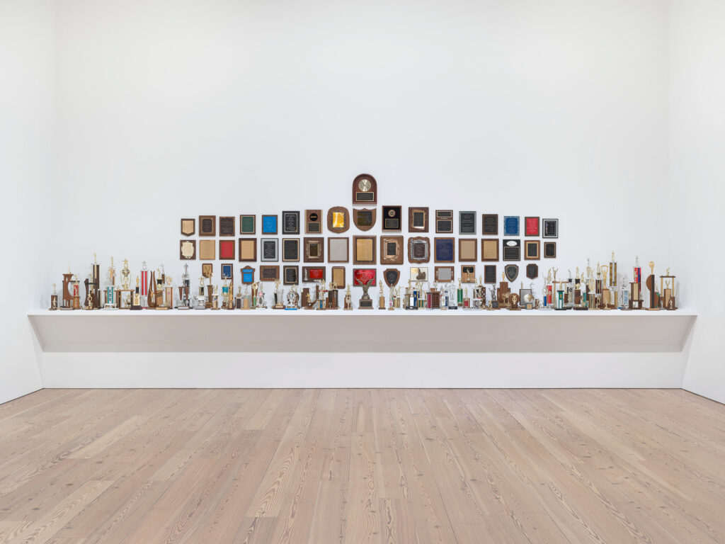 An art installation in a room with all white walls. The installation displays various types of trophies sitting on a large wall-to-wall shelf and plaques mounted on the wall.