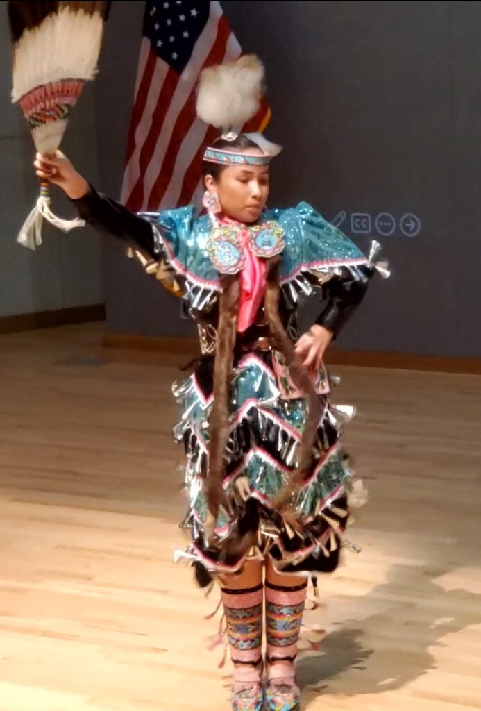 A performer wearing a traditional Native American jingle dress dancing with one hand raised and another at their hip.