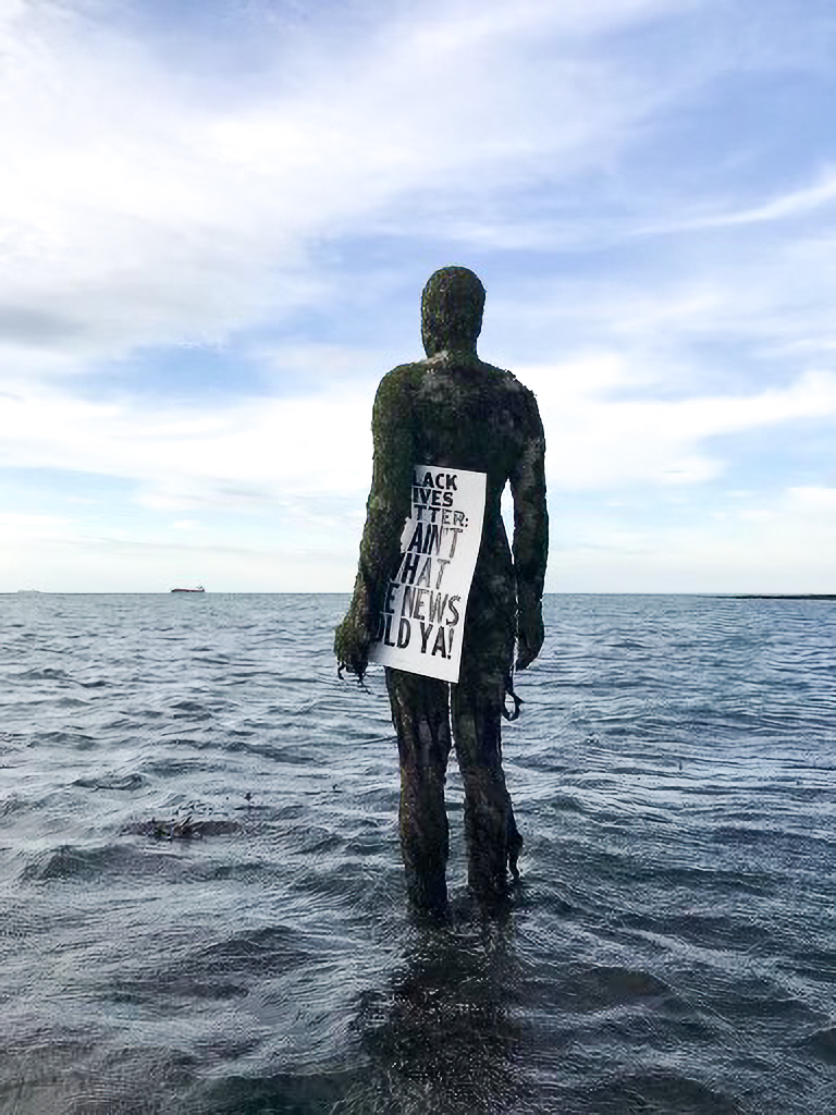 A stone sculpture of a standing human figure with the legs submerged in a large water body is overgrown with moss. It is looking out to the expanse of the water as it holds a letterpress poster that reads "Black Lives Matter: It ain't what the news told ya!" in bold blank all-caps type.