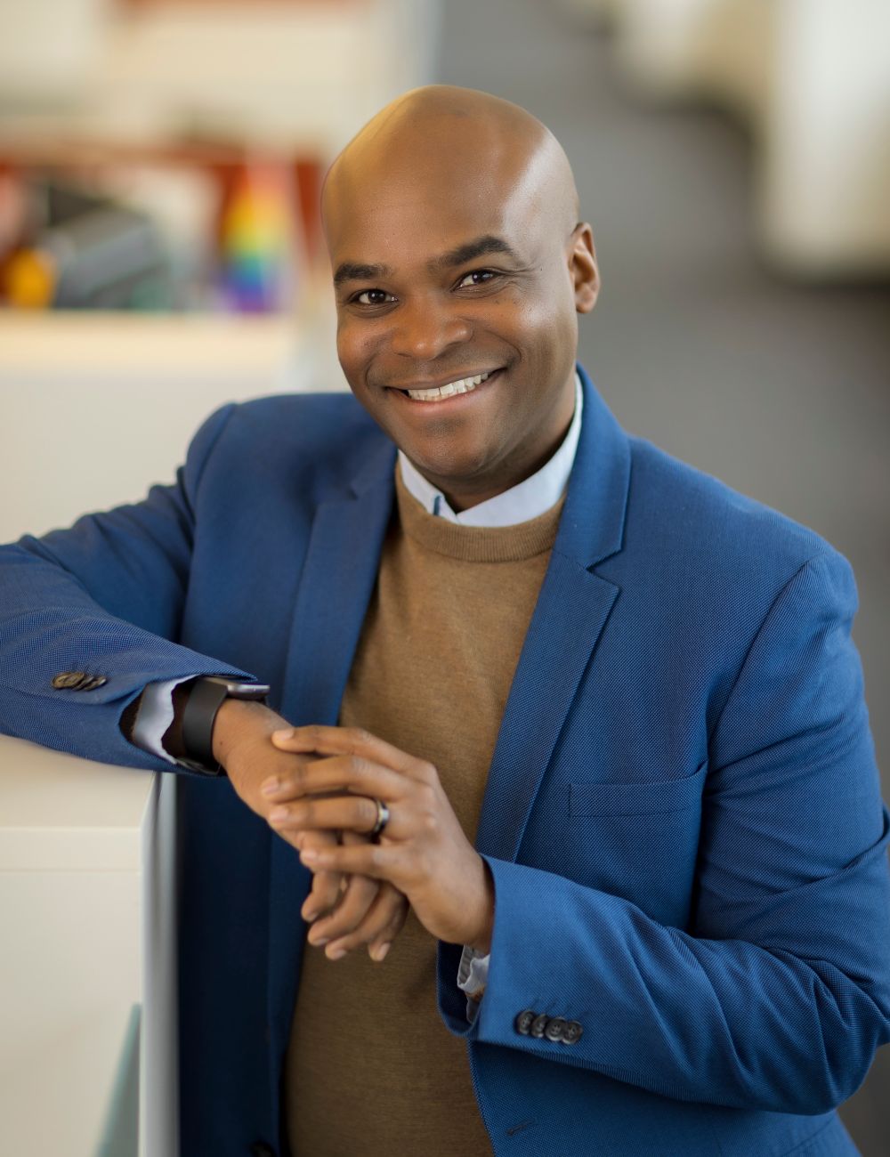 Headshot of a smiling person of medium dark skin tone, who is bald and wearing a brown sweater under a blue suit jacket, with hands crossed and leaning against a cubicle.