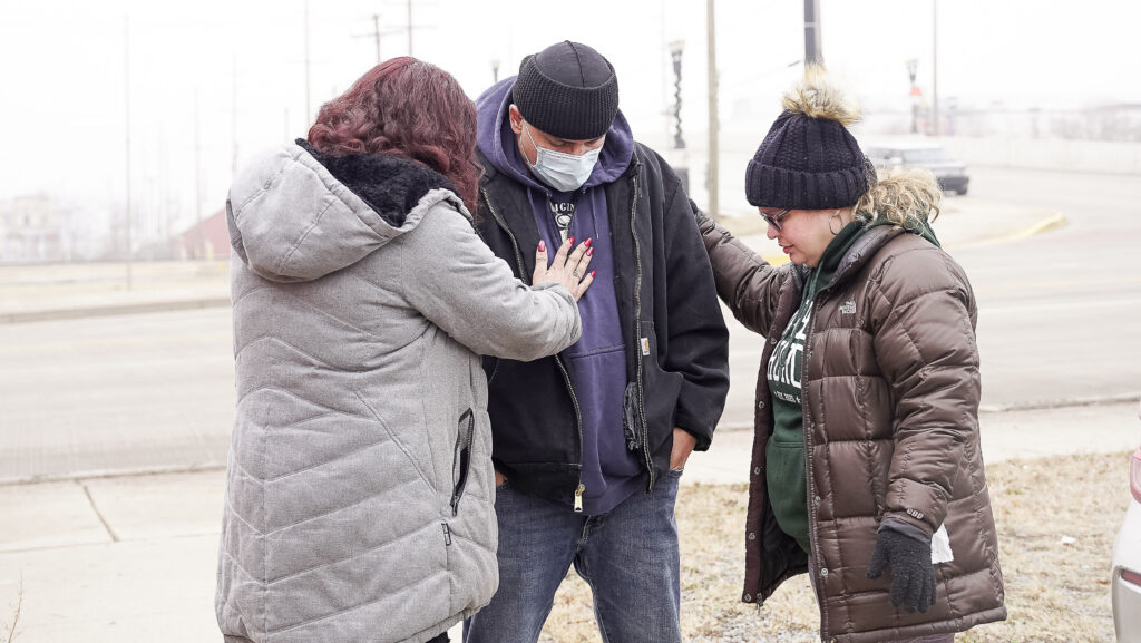 Two people wearing winter coats stand by and put their arms around a person wearing a face mask and sweatshirt.