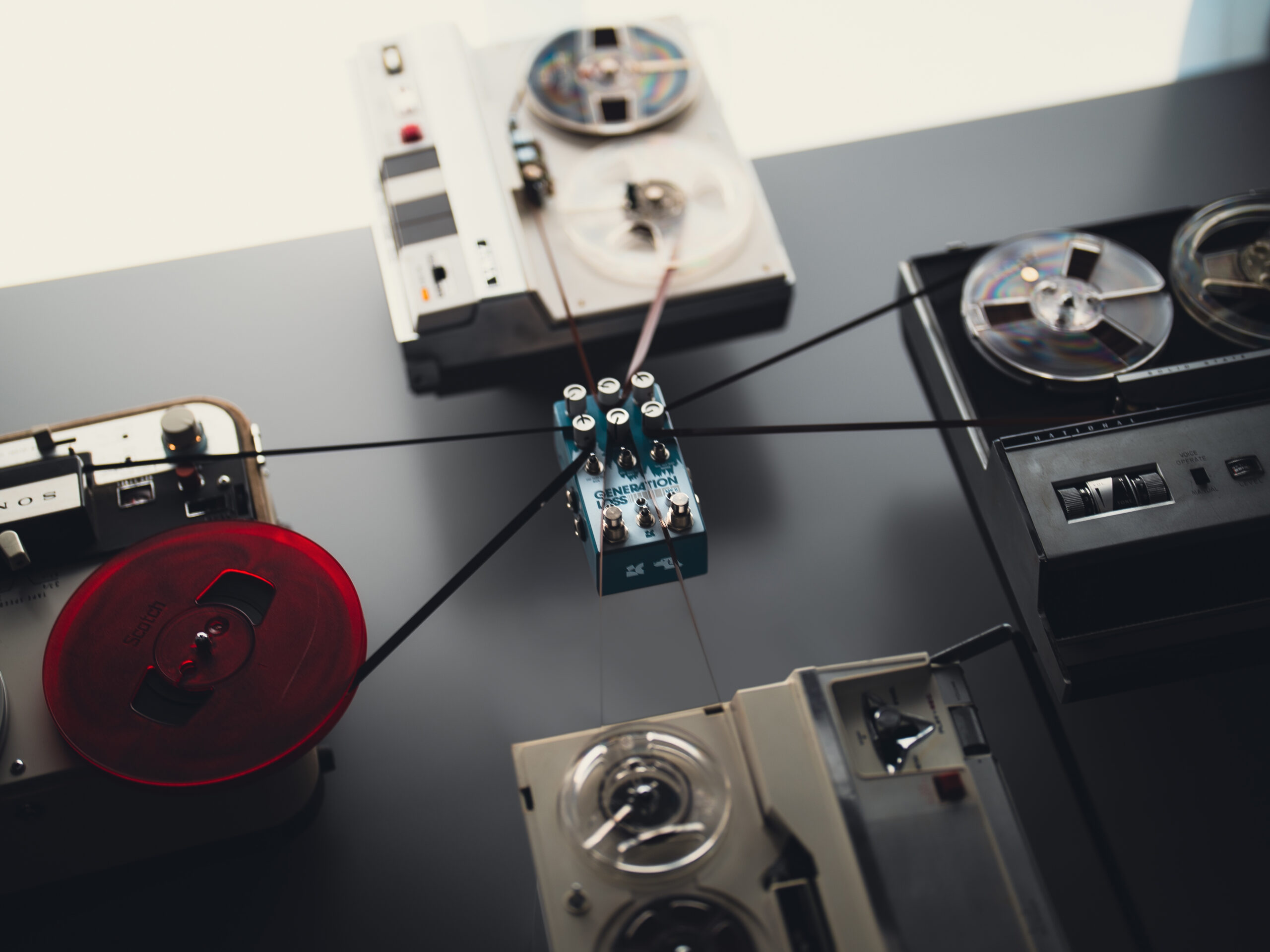 A product photo depicting a blue guitar pedal strung up by the tape of four different vintage recorders.