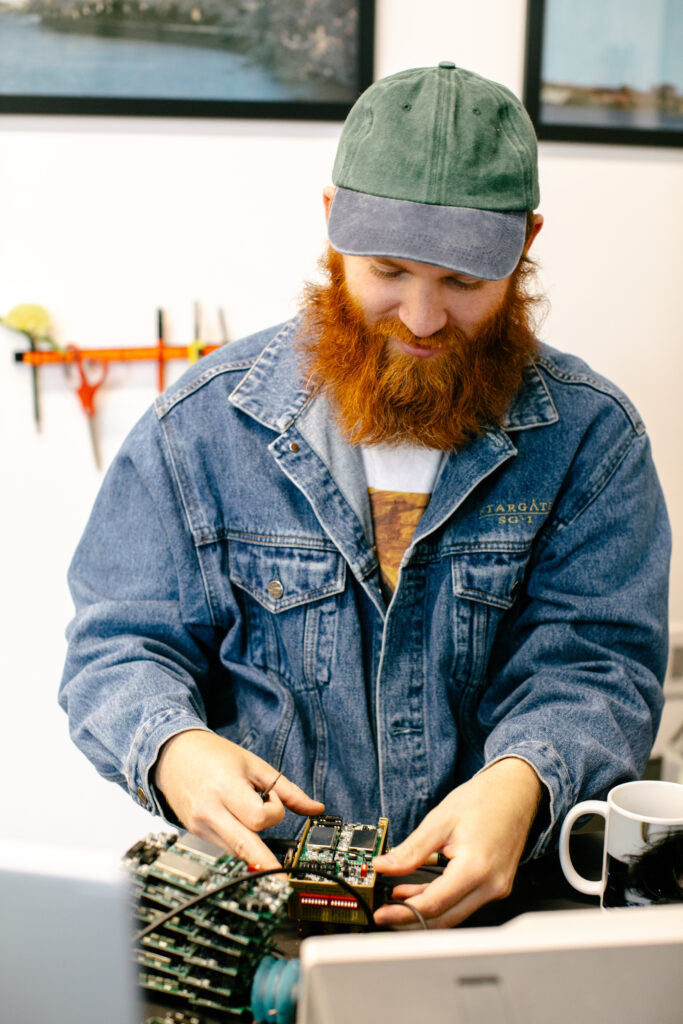 A person of light skin tone with a thick long orange beard looks down as they wire and solder a circuit board of a guitar pedal. They are wearing a faded green and grey baseball hat and a blue denim jacket.