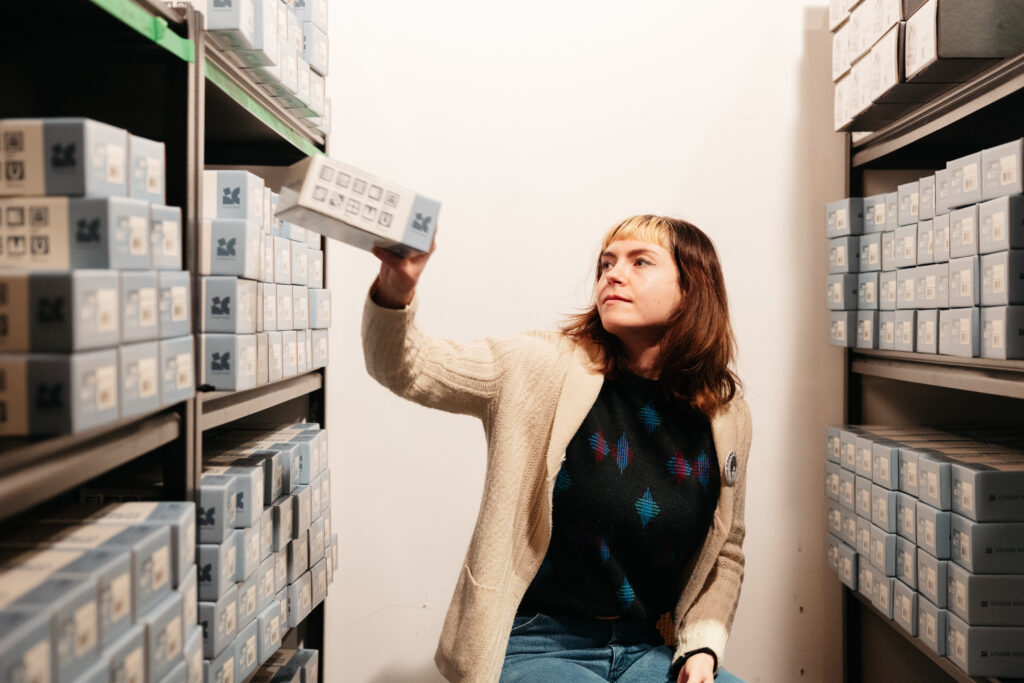 A person of light skin tone wearing a dark top, jeans, and a beige cardigan, is sitting and in the middle of putting away a box on a shelf. They are in between two rows of shelving, and they are filled with similar boxes as the one she is putting away.