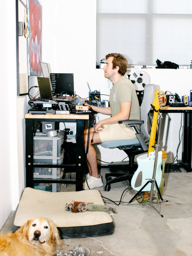 A side view of a person of light skin tone with medium brown hair as they sit in a chair and face multiple computer screens. In view are other electronic objects, including a light blue electric guitar on a stand. In the foreground, almost out of focus, is a midsize golden-haired dog laying next to a dog bed on the floor.