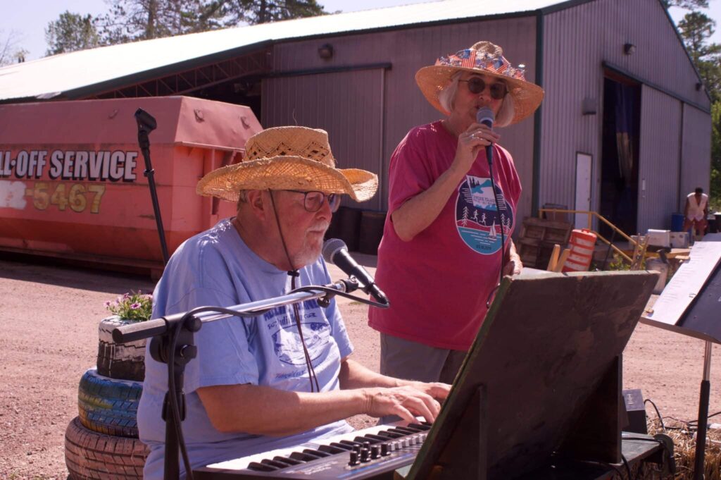 Two older people in straw hats sing into microphones on a sunny day outdoors at a recycling plant