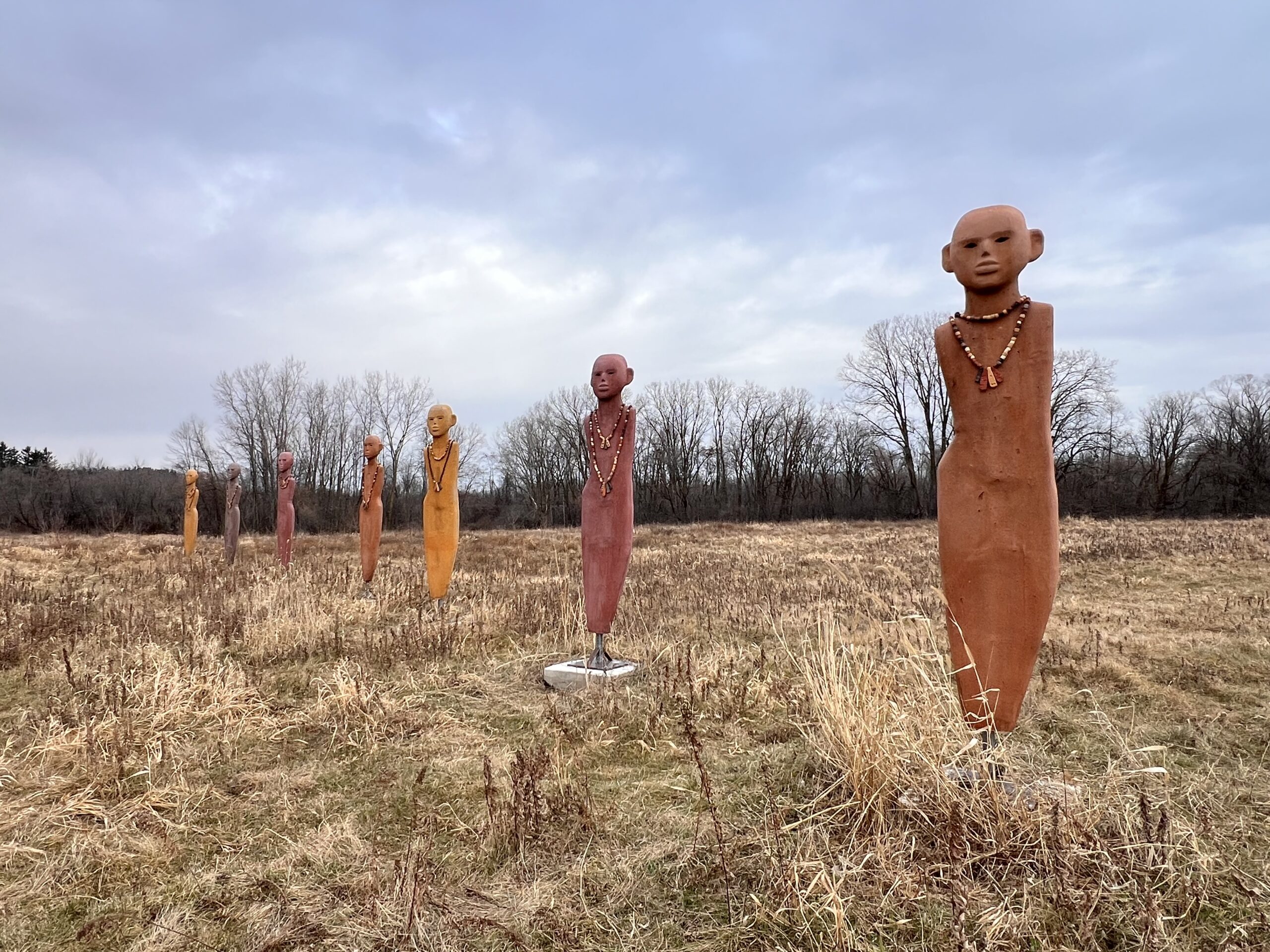 An outdoor field with seven figurative sculptural works that look like abstracted human forms in shades of reddish browns and yellows.