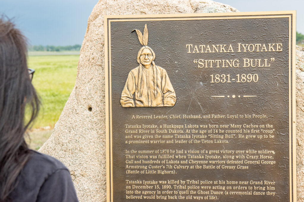 A dark metal plaque with a gold-colored border and text. It has an image of a person in traditional Native American headgear with two feathers. The title of the plaque reads 'Tatanka Iyotake Sitting Bull 1831-1890.'