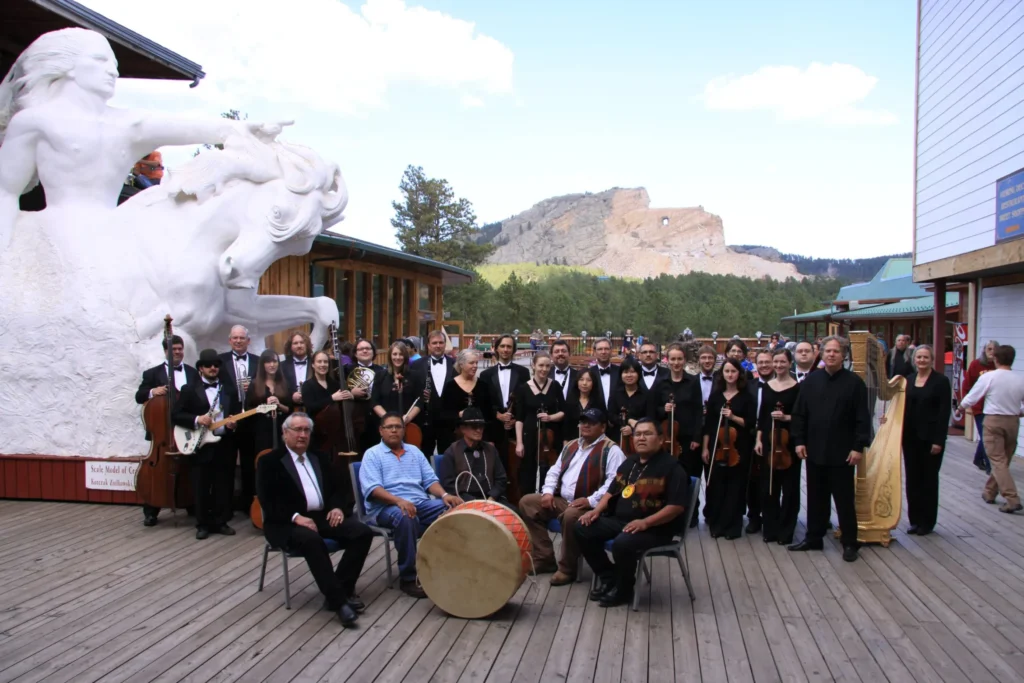 Twenty-seven orchestra members in formal dress stand with instruments behind a group of five men who sit in front of a large traditional drum. They are outside on a sunny day on patio in front of the Crazy Horse sculpture in South Dakota.