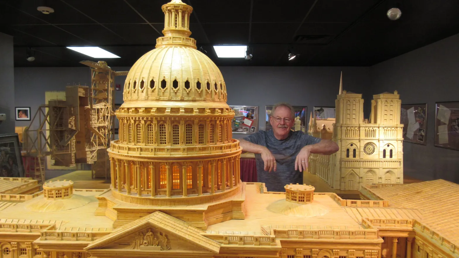 An older person poses with elbows resting on a glass barrier surrounding a model of the U.S. capitol that appears two heads taller than them.