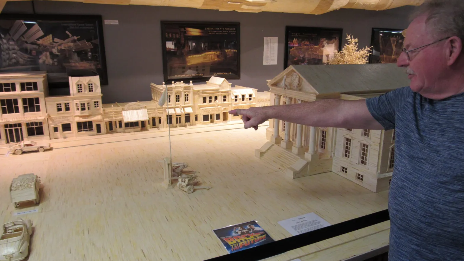An older man in a blue shirt points at a miniature cityscape constructed of matchsticks.