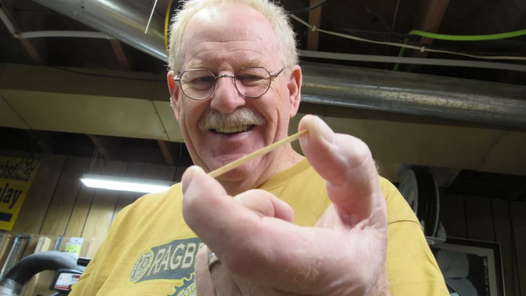 A smiling older man in a bright yellow shirt holds a matchstick close to the camera between his thumb and pointer finger.