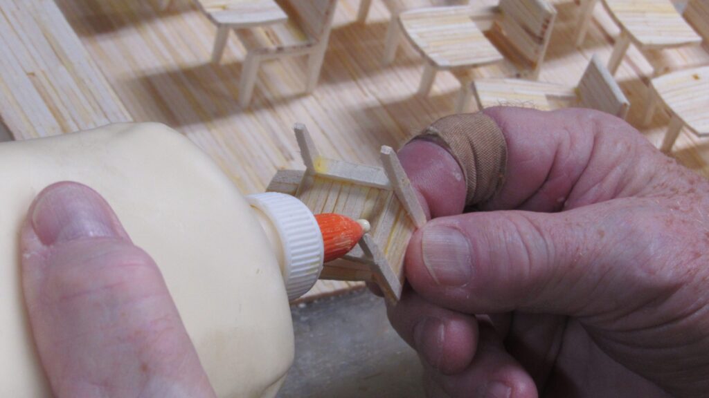 A pair of hands holding a bottle of glue and a miniature chair made of matchsticks.