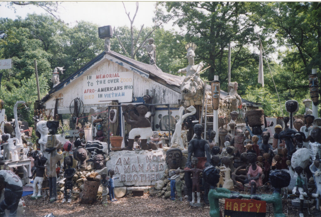 A white wooden house surrounded by sculptural works depicting figures in different poses. There is lettering on the house that reads "in memorial to the over 7,000 Afro-Americans who died in Vietnam."