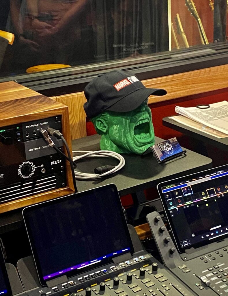 A recording studio soundboard with multiple laptops and interfaces, with a green Hulk head with a Marvel Studios hat sitting on a shelf.