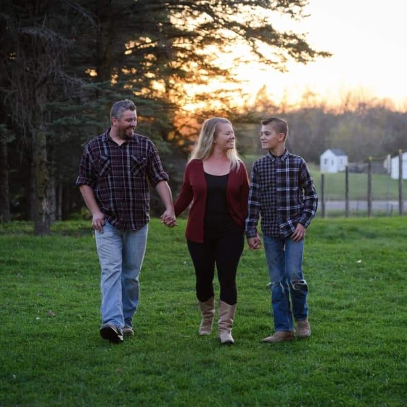 Two adults and a young adult hold hands and walk on green grass. The sun is setting behind them.