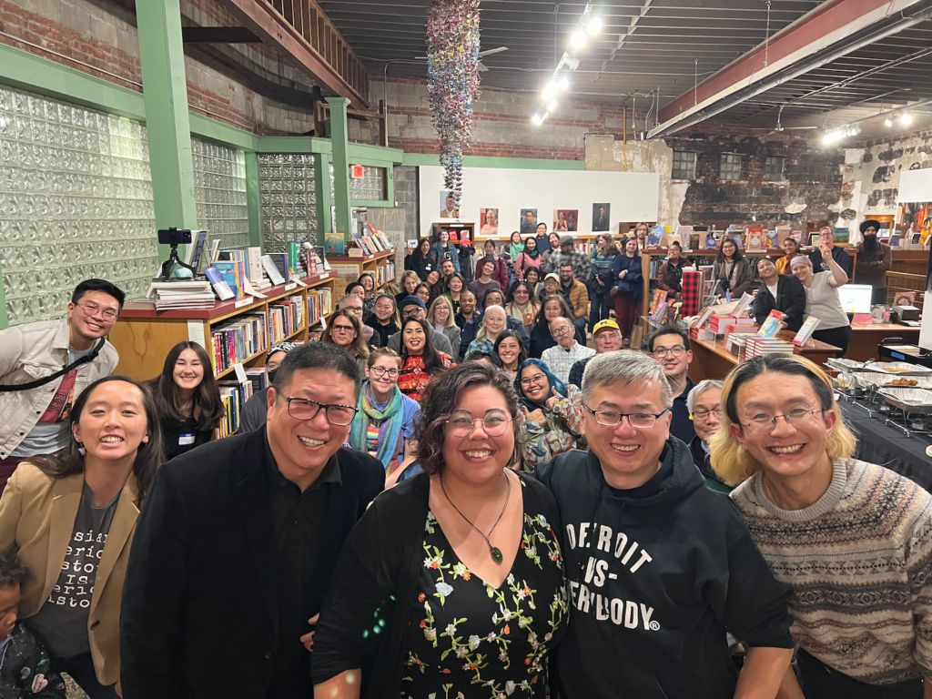 A large group of smiling people look at the camera from between the shelves of a spacious high-ceilinged bookstore.