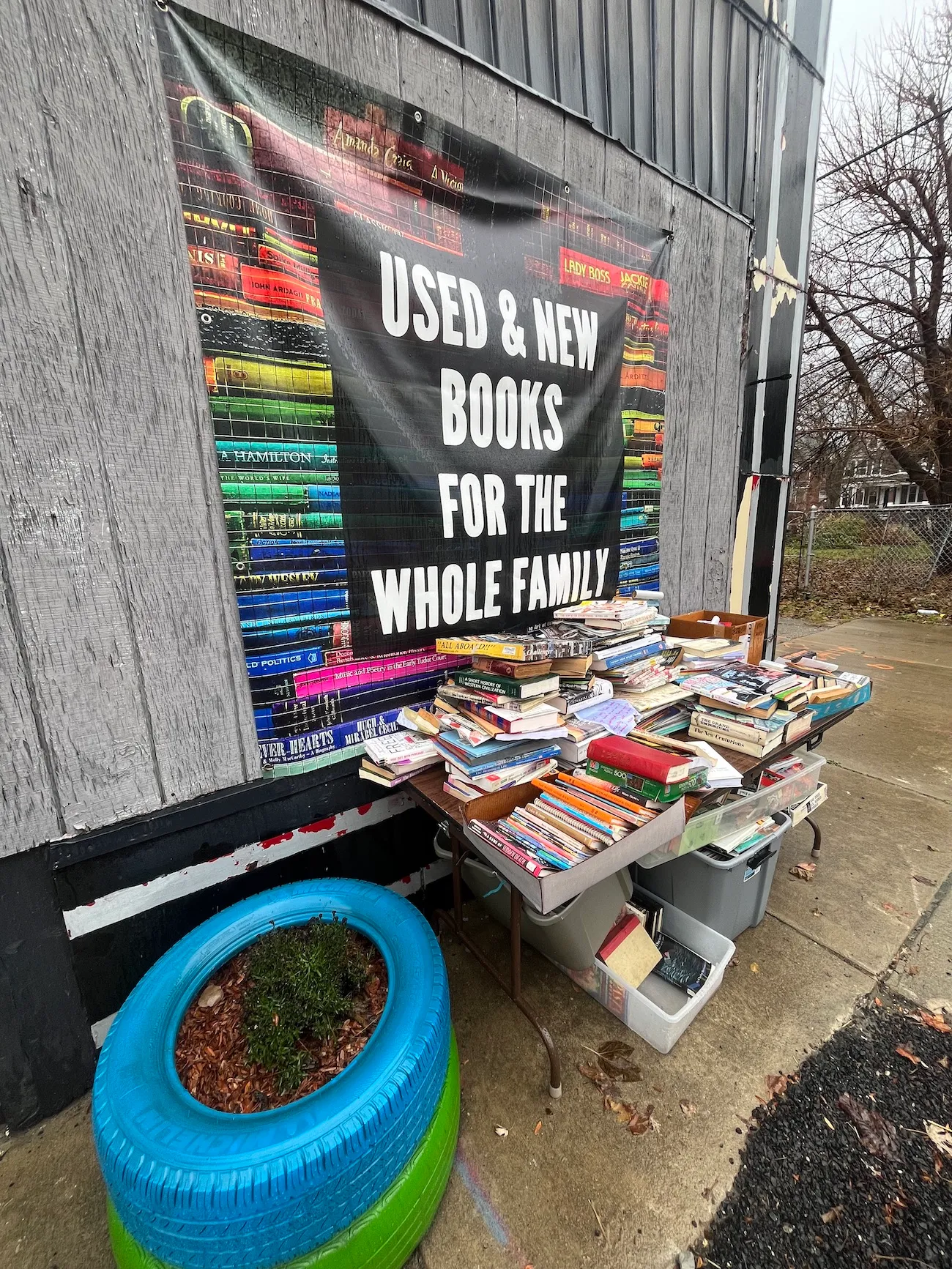A stack of books on a table outside underneath a colorful banner reading "Used & New Books For The Whole Family." Two stacked car tires, one blue and one green, converted into a planter are in the foreground.