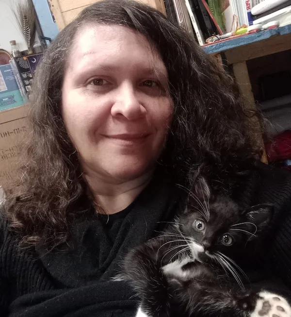 A person in a black shirt and long curly brown hair holds a small black and white cat staring into the camera.