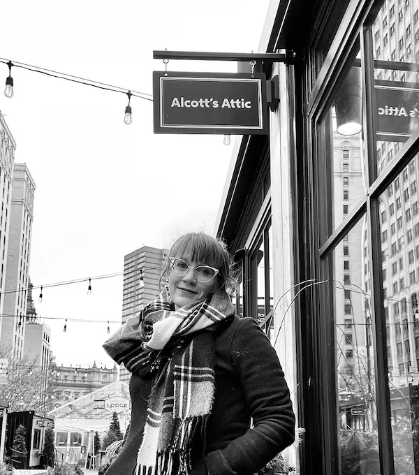 A black and white image of a person with bangs and glasses and a winter coat and plaid scarf below a sign reading "Alcott's Attic" on a city street with string lights overhead.