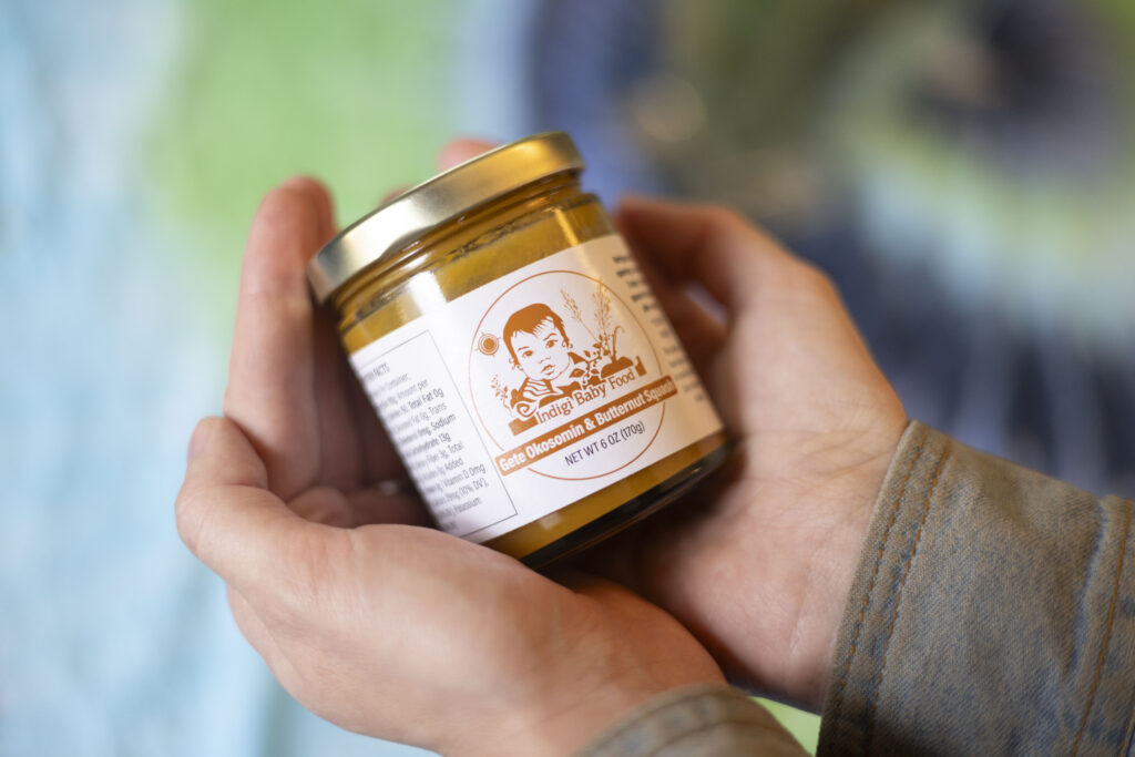 A pair of cupped hands hold a glass jar of mustard-colored baby food. The label on the jar has a drawing of a baby and plants. It reads "Indigi Baby Food Gete Okosomin and Butternut Squash"