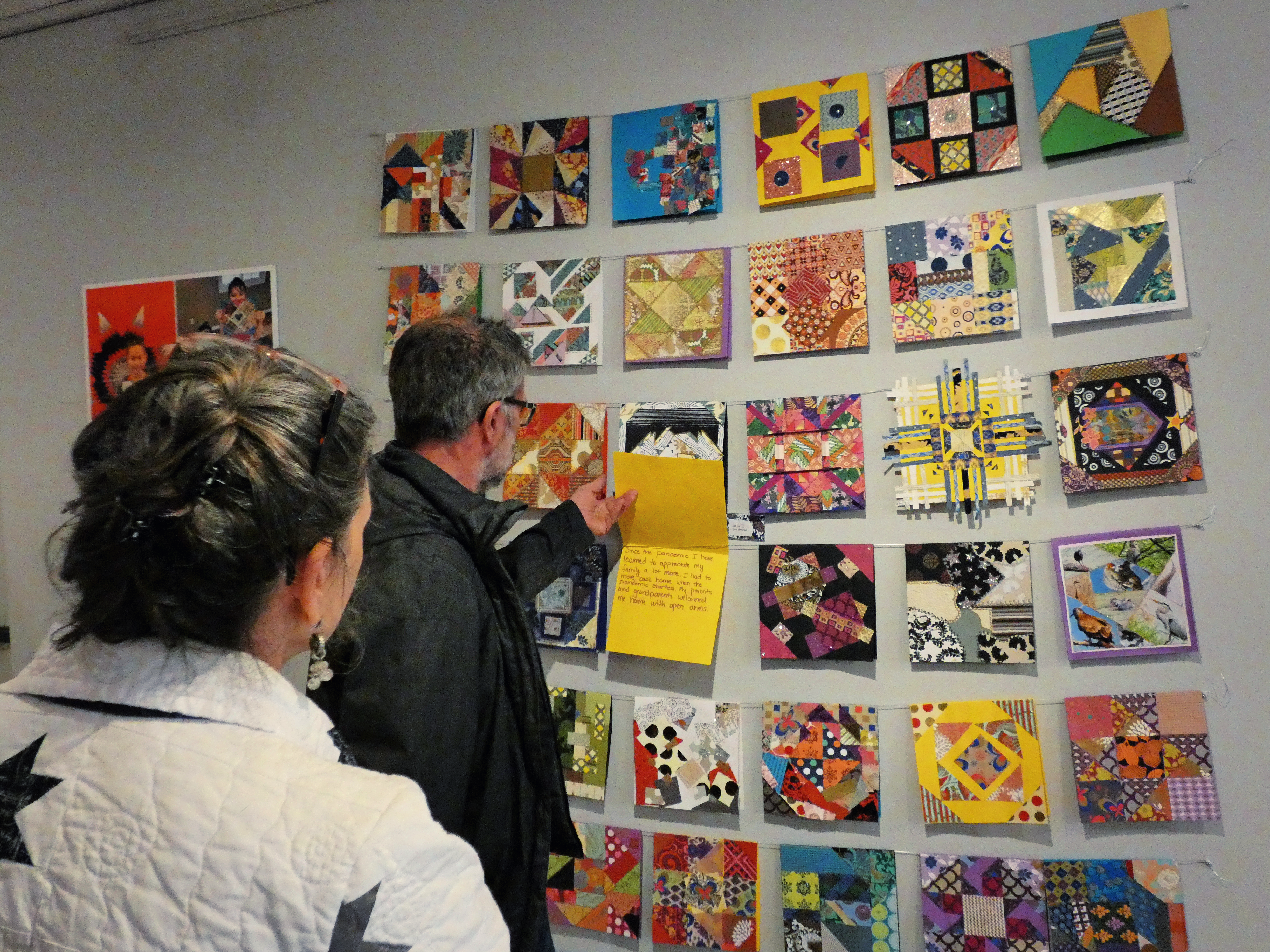 A person holds open an artwork mounted on a gallery wall. It is a part of an installation with other similar card-like colorful and patterned works of paper. They are all strung by thread in rows.