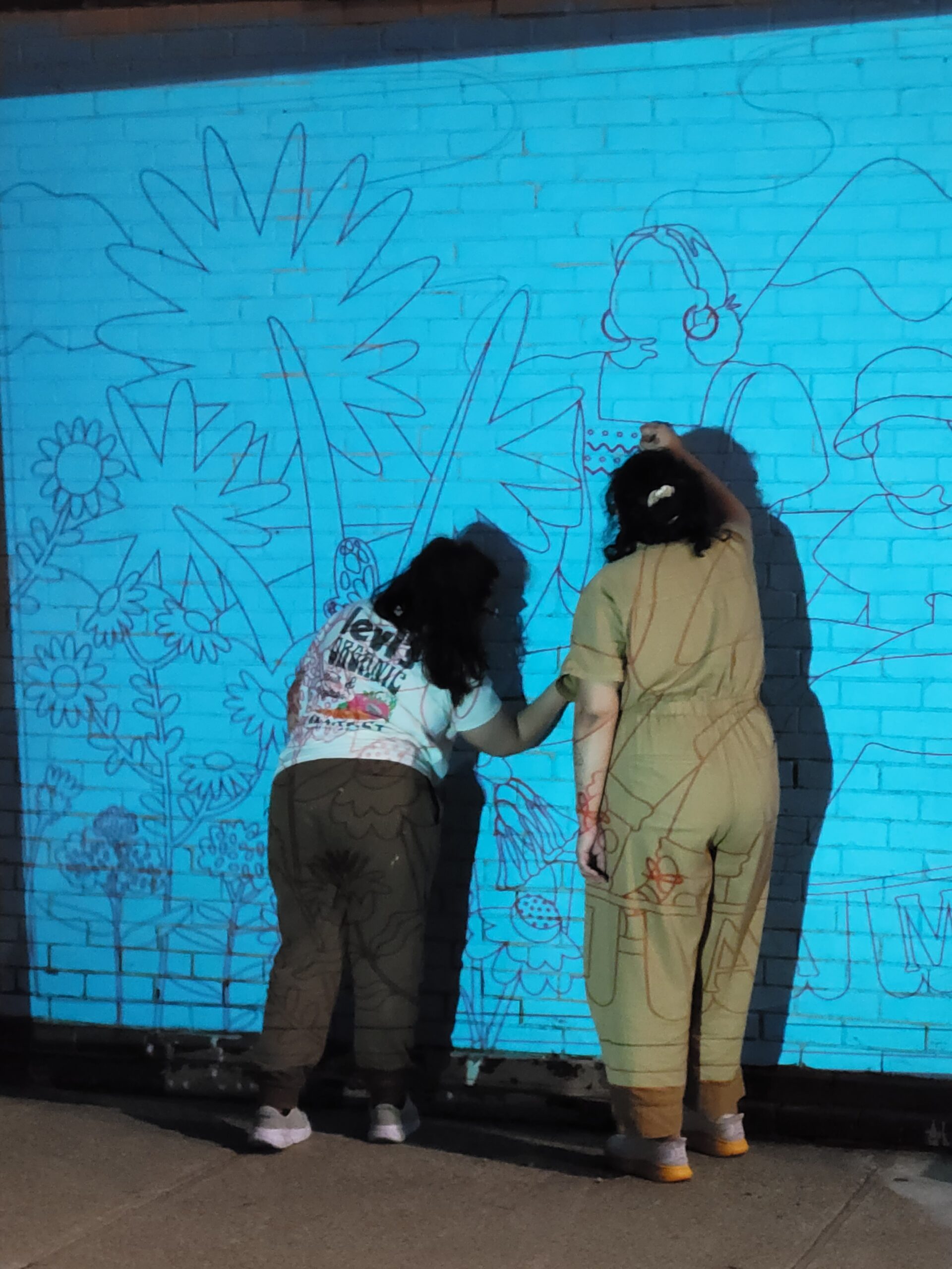 Two people sketching outlines of flowers and people on a blue wall.