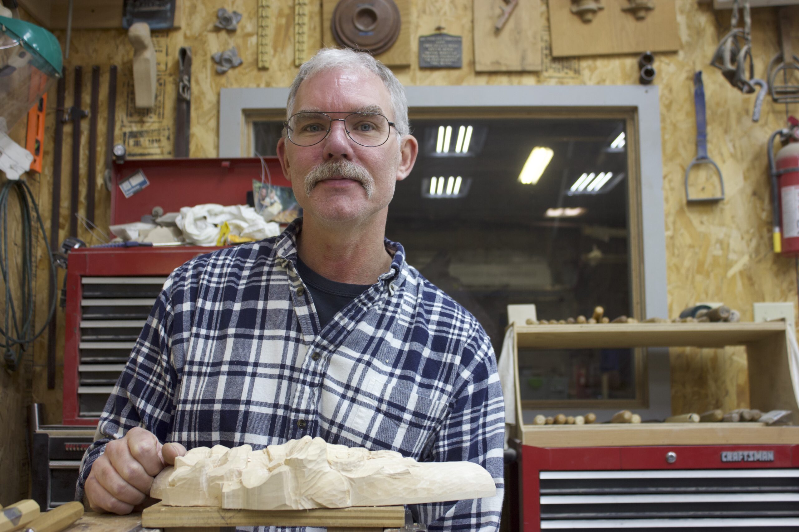 A photo of an older man with light skin tone, glasses, and a mustache in a wood shop