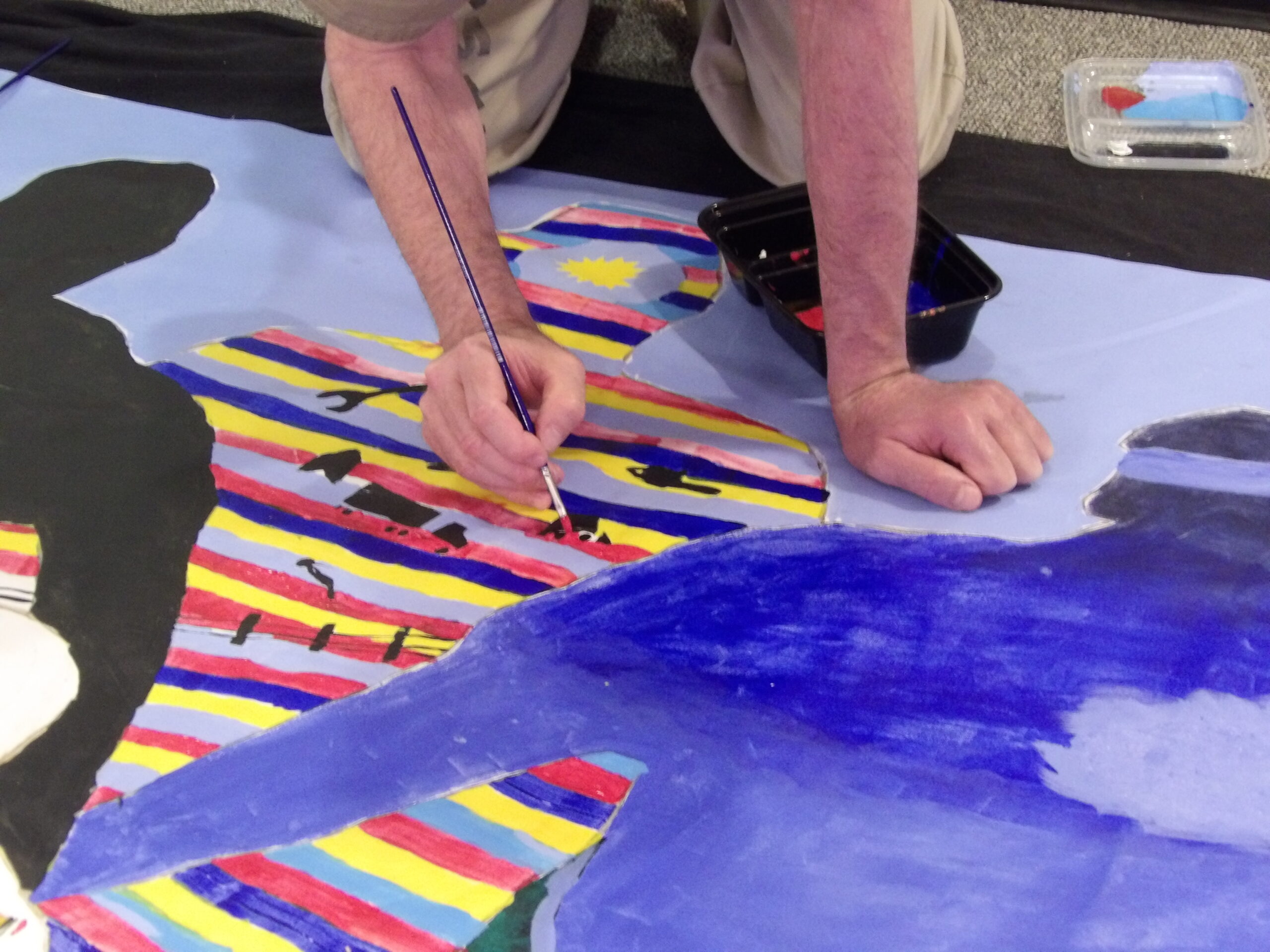 An in-progress painting with colorful figurative silhouettes rest on the ground as a person holding a paint brush works on it.