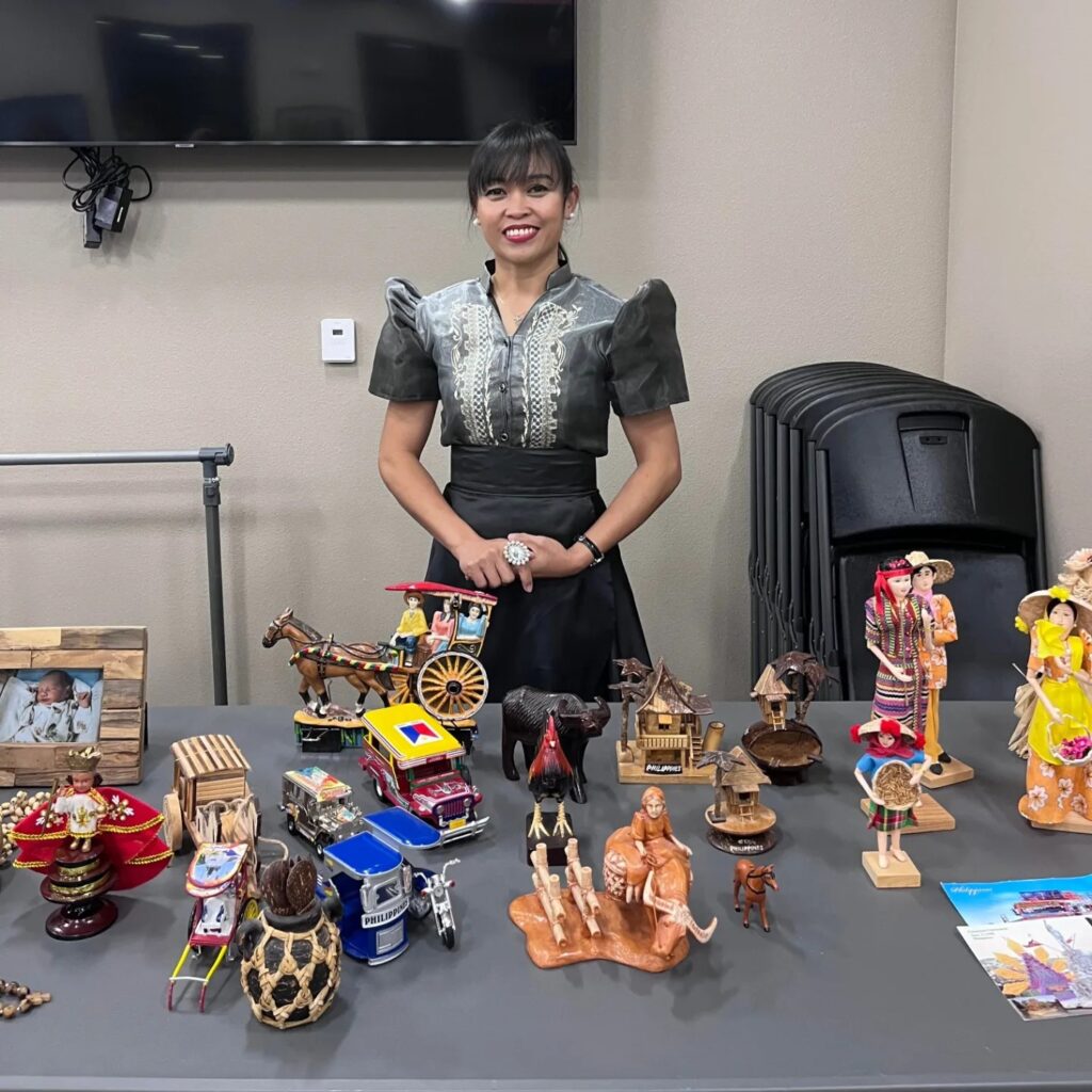 A person of medium skin tone dressed in a black dress stands behind a table and smiles. On the table in front of them are different sculptural objects.