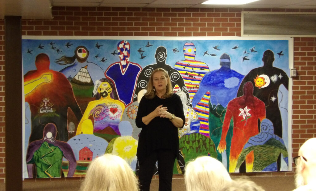 A person of light skin tone and dark blonde hair speaks to an audience. They are wearing a black top and pants, and there is a large mural painting behind them.